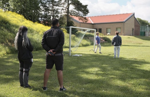 Två ungdomar spelar fotboll medan två medarbetare tittar på från sidan.