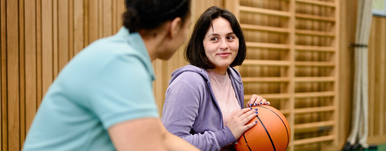 En medarbetare och en ungdom med en basketboll i knäet samtalar på en bänk i en idrottshall.
