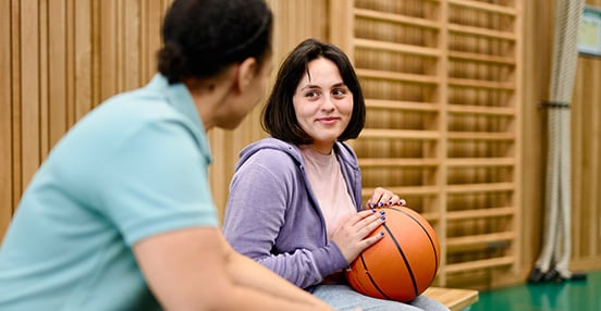En medarbetare och en ungdom med en basketboll i knäet samtalar på en bänk i en idrottshall.