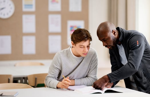 En pojke som sitter i ett klassrum med skolböcker och får hjälp av en lärare.