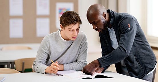 En pojke som sitter i ett klassrum med skolböcker och får hjälp av en lärare.