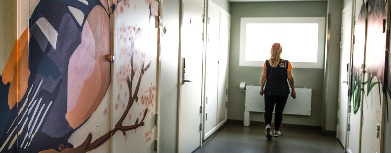 Bild visar en kvinna i arbetskläder i en korridor med stängda dörrar.