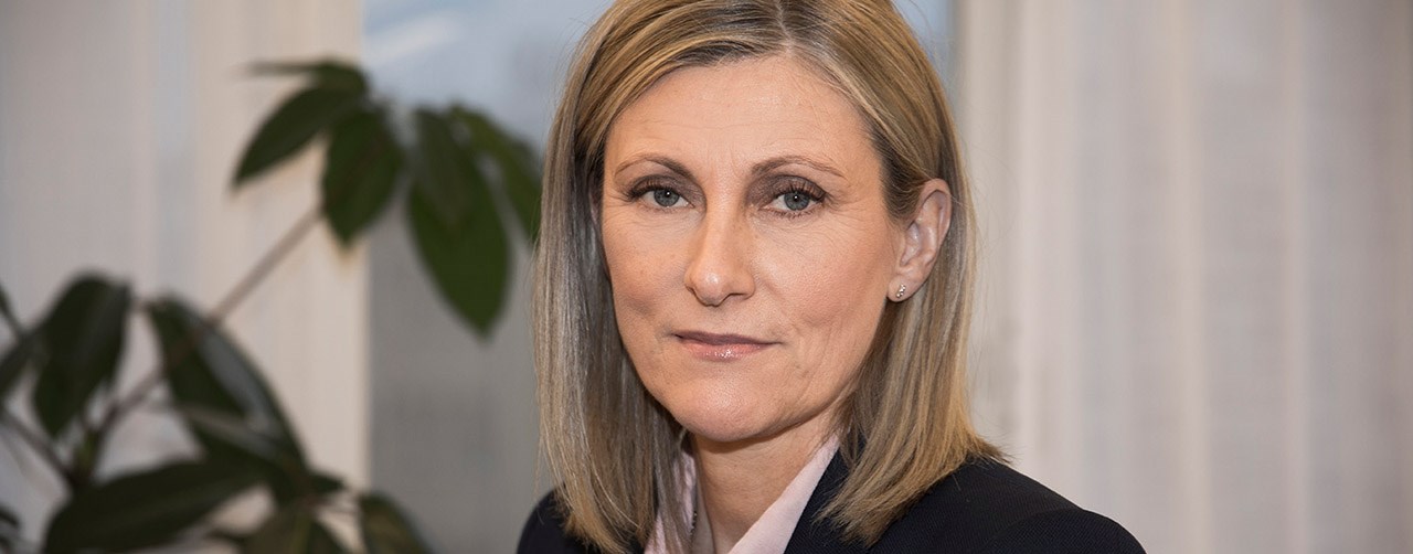 Bilden visar en kvinna med seriöst ansiktsuttryck. I bakgrunden syns delar av en grön växt samt ett fönster. 